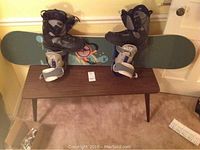 Full view of the snowboard laid on a table with attached bindings and boots placed on top, plus visible stamp pad nearby.