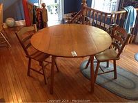 Round solid maple drop leaf table with two leaves extended and two chairs placed alongside on wooden floor.