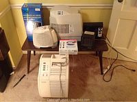 Wide view showing several air purifiers and heaters on and around a small table, including the 3Q purifier in box, Honeywell purifier, and smaller heaters.