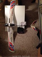 Top down image showing the full RC plane with the remote and manual placed beside it on a carpeted floor. The plane has red, blue, and black logo graphics and a wingspan of about 50 inches.