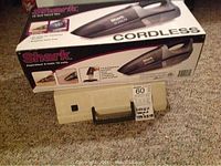 Box of Shark 12V cordless vacuum cleaner with label and beige Olympia typewriter case below it on carpeted floor.