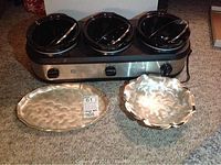 Full view of the TRU 3-pot slow cooker with stainless steel base and three black pots with lids, alongside two hammered WMF-Ikora stainless steel serving trays, one oval and one round with decorative edge.