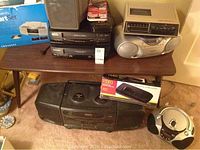 Photo showing table with stacked VCRs, karaoke machine, alarm clock CD player, and boombox on floor with headphones.