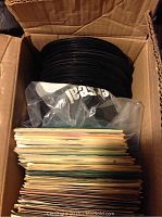 Stack of over 100 black vinyl records stacked inside an open cardboard box with several paper sleeves visible.