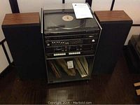 Full frontal view of the Lloyds stereo system with turntable on top, amplifier and cassette unit below, black speakers with wood paneling on sides, stereo cabinet with vinyl records on lower shelf.
