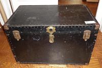 Front side view of large black steamer trunk showing lock and latches, metal studs and corner protectors, and wear on surface.