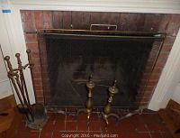 Photo of the fireplace area showing the brass mesh screen, fireplace tools in the stand, and andirons in front of the fireplace.
