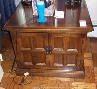 Front view of square wooden side table showing paneled cabinet doors, polished finish, and metal handles.