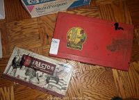 Top view of two vintage Erector sets: one red metal box with worn graphics and a sticker, and one cardboard box with Erector branding showing some metal parts inside.
