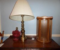 Photo showing the vintage brass ram's head table lamp with a beige fabric shade beside the wooden glass front display shelf on a wooden surface against a light blue wall.