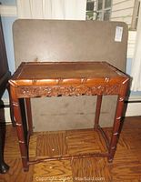 Small carved wooden side table with floral carved apron and faux bamboo legs, placed on wooden floor in front of a vinyl-covered board.