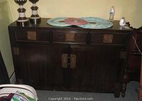 Front view of dark wooden credenza with three drawers and two doors with brass hardware, sitting against wall with items on top