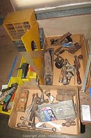 Wide view showing multiple vintage hand tools and hardware organized in trays and boxes. Items include hand drills, wrenches, screwdrivers, saws, and other woodworking tools.