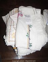 Stack of folded embroidered table linens with visible needlepoint and crewel floral designs and lace edges.