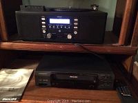 Photo showing black TEAC Lap-R450 turntable, cassette, and CD combo unit on upper shelf and Philips VHS player with remote and manual on lower shelf in wooden shelving.