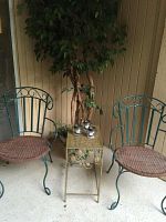 Photo showing the two green metal outdoor chairs with red cushioned seats, a small decorative gold-toned metal side table, an artificial ficus tree behind the chairs, and four silver tea light holders placed on the table.