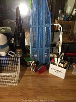 Photo showing blue metal ironing board, upright Eureka vacuum cleaner, handheld dust buster vacuum, black step stool, and housekeeping box on wooden floor.