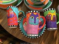 3 ceramic mugs and 4 plates displayed on a wooden table, each with a colorful artistic cat face design and checkerboard border patterns.