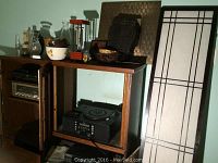 Photo showing a 3 panel Asian screen with white and black linear design leaning against wall. Various decorative vases in clear and white ceramic with floral patterns on a wooden shelving unit. Black and natural woven baskets and a small mirror with textured frame are visible along with a clear glass oil lamp.
