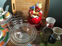 Photo of ceramic cookie jar, clear drinking glasses, Pyrex bowls, assorted mugs, and colorful plates on a surface.