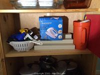 Photo showing a wooden shelf containing a box with Toastmaster 5-speed hand mixer, white plastic colander with small kitchen utensils inside, orange plastic jug with lid, spice display rack with spice containers, metal pots with glass lids, and some small kitchen gadgets including a blue and yellow juicer and a thermometer.