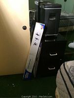 Photo shows black paper shredder, black 2 drawer file cabinet with silver handles, boxed easel, and loose white paper.
