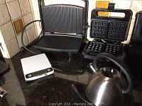 Photo showing Salton Belgian waffle maker with removable waffle plates, Breville grill or panini maker with ridged plates, stainless steel kettle, and digital food scale on countertop.