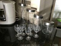 Photo showing four glass canisters with silver metal lids alongside six clear stemmed aperitif glasses with etched designs.
