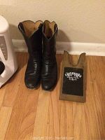 Pair of black leather Justin cowboy boots size 6.5 D shown standing side by side on wooden floor next to a wooden Cavenders boot jack, all in good condition with light wear.
