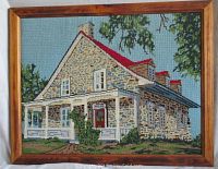 Framed needlepoint showing a farmhouse with stone walls and a red roof set against a blue background sky.