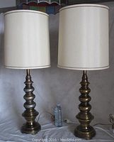 Pair of tall table lamps with burnished brass finish bases and cream-colored fabric shades side by side, shown plugged in and powered off with water bottle for size reference.