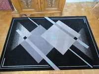 Top-down photo of the rectangular Eaton European Collection area rug with geometric gray and rose patterns on black background, showing most of the rug and some surrounding furniture.