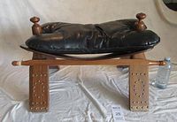 Side view showing the wood camel saddle foot stool with black leatherette cushion, studs on legs, and two side knobs.