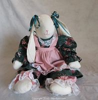 Front view of the handcrafted stuffed rabbit toy sitting upright, showing long ears tied with teal ribbons, facial markings, and dressed in floral dress with pink pinafore and lace trim.