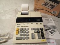Canon P102-DIII desktop calculator showing front view with paper roll installed, dust cover, extra roll holders, user manual visible in background
