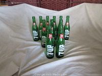 Eleven vintage green glass 7Up bottles arranged in a pyramid shape on a white patterned surface under indoor lighting.