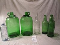 Five glass bottles including two large green jugs and three smaller bottles on a white backdrop with a water bottle for scale.