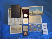 Overview photo of the four coin sets including a black display case with coin slots, a proof silver dollar in a red velvet presentation box, informational cards, and a sealed plastic holder with coins.
