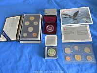 Photo showing four coin sets including uncirculated and specimen sets in plastic cases and two silver dollar coins in presentation boxes with informational inserts visible around them.