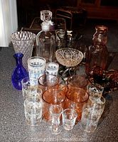 Glassware set displaying decanters, vases, pink mugs, multiple shot glasses, and colored glass pieces on kitchen counter.