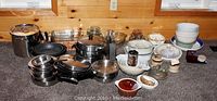 Wide shot showing entire assortment of stainless steel pots, nonstick pans, glass baking dishes, bowls, and serving dishes arranged on floor and a wooden surface.