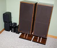 Photo of a pair of large PSB70R floor-standing speakers with wooden stands and black grilles, alongside several smaller speakers and units on a carpeted floor in a corner.