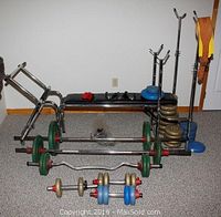 Wide view of weight bench, bars with cast iron, plastic filled York and Weider weights, dumbbell bars, collars, and leather belt on floor.