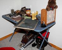 Overview of the collectibles items arranged on a blue folding table including ledger, toy bear, cameras, tin container, keys, and kitchen items.