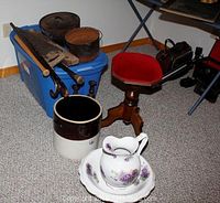 Two cast iron pots on blue plastic tote, piano stool with red cushioned seat nearby, crock, wash set with bowl and pitcher in front, old levels, tools, canes, and bat visible