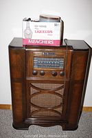 Front view of large wooden GE console radio with tuning dial, control buttons, and decorative speaker grille. Box of old tubes on top.