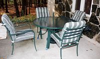 Overall view of round metal patio table with green textured glass top and four cushioned matching chairs positioned around it near stone wall.