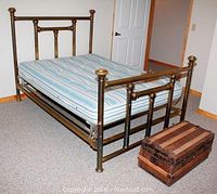 Full view of brass double bed with striped mattress and wooden trunk placed in front