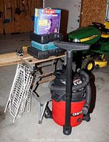 Photo showing red Craftsman wet/dry vacuum with hose, Workmate bench with the drill cases and fire escape ladder box on top, and white metal shoe rack beside it.
