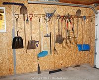 Wide photo showing an assortment of yard tools hanging on a wall in a garage. Items including shovels, rakes, brooms, and clippers are visible.